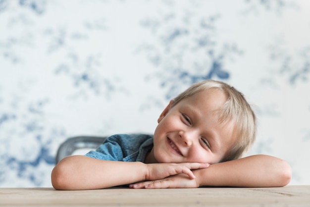 Niño pequeño sonriente que inclina su cabeza en mano sobre la mesa de madera