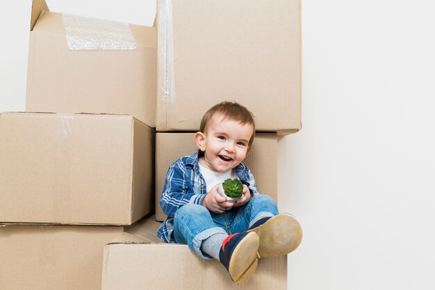 Niño pequeño sonriente que se sienta en la caja de cartón móvil que sostiene la planta del cactus