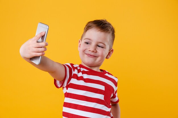 Niño pequeño sonriente que sostiene el teléfono móvil y que hace selfie