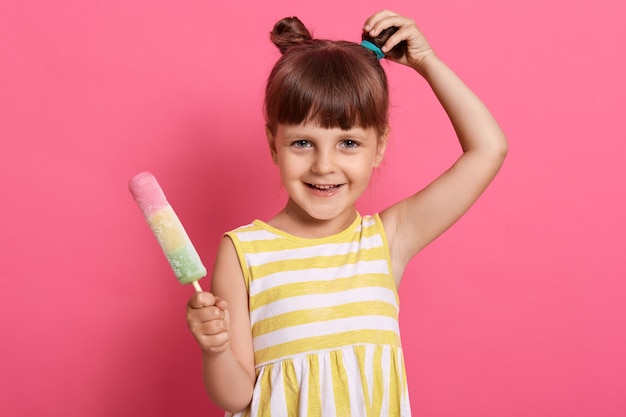 Niño pequeño con sonrisa encantadora, moño de pelo, vestido de sarafan rayado amarillo y blanco, mira a la cámara, posa aislado sobre fondo rosa, tocando su nudo.