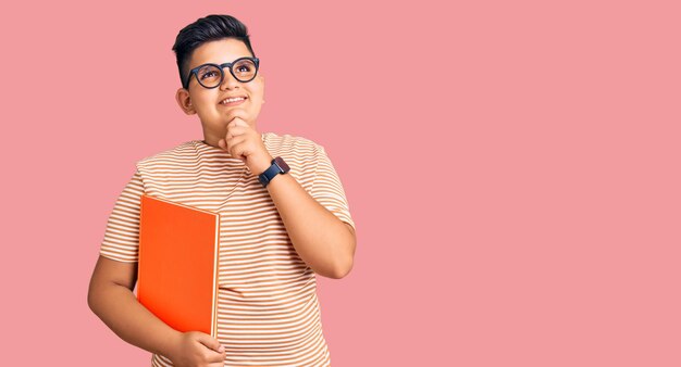 Niño pequeño sosteniendo un libro con gafas con la mano en la barbilla pensando en la pregunta expresión pensativa sonriendo y pensativo cara concepto de duda