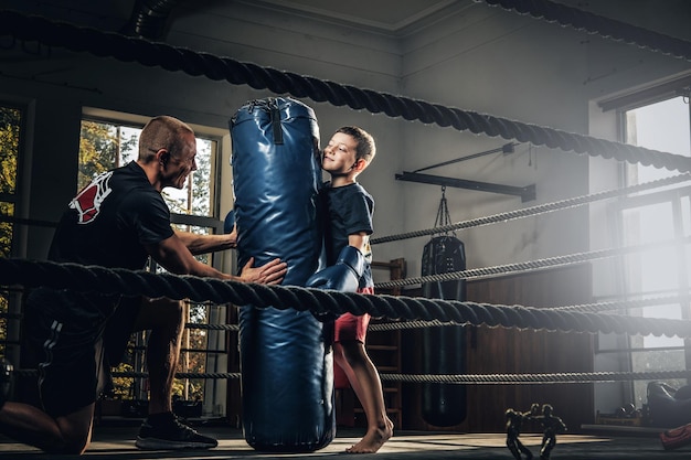 El niño pequeño tiene un entrenamiento de boxeo serio con entrenador y saco de boxeo.