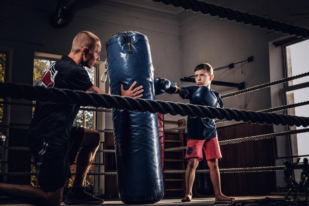 El niño pequeño tiene un entrenamiento de boxeo serio con entrenador y saco de boxeo.