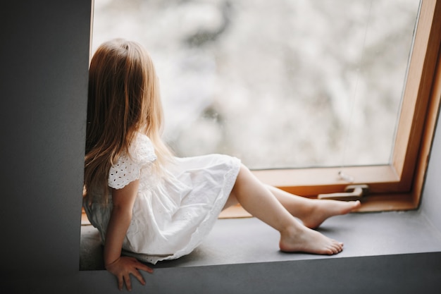 Niño pequeño en tierno vestido blanco está sentado en el alféizar de la ventana