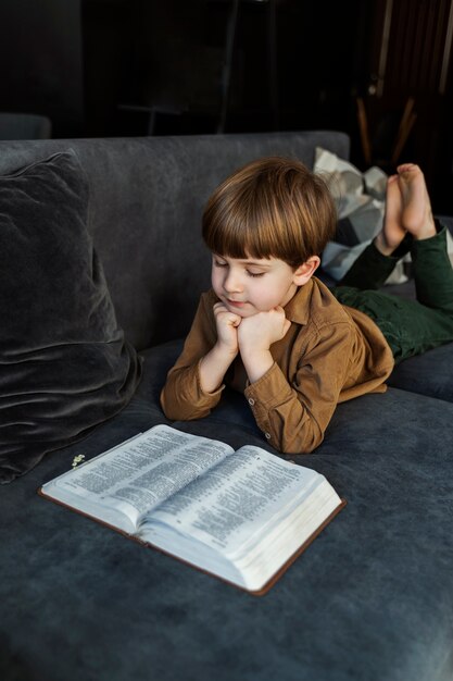 Niño pequeño de tiro completo leyendo la biblia en el sofá