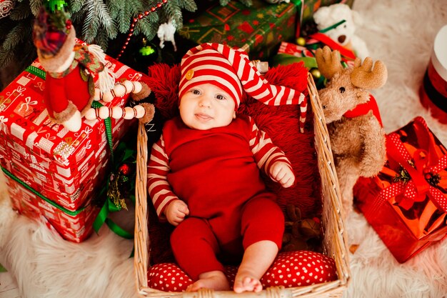 El niño pequeño en traje rojo divertido y sombrero largo miente en la cesta antes de un árbol de navidad