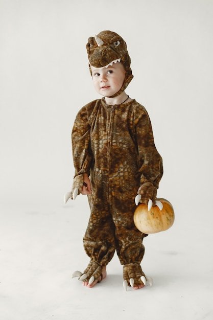 Foto gratuita niño pequeño vestido con traje marrón de un dinosaurio sosteniendo una calabaza. el niño tiene una capucha con cara de dinosaurio.