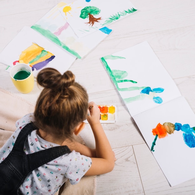 Niño pintando con acuarelas sobre papel y tendido en el suelo.