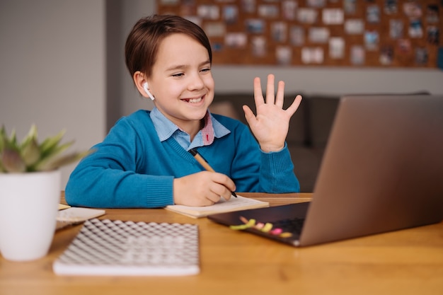 Un niño preadolescente usa una computadora portátil para hacer clases en línea, saludando a la maestra