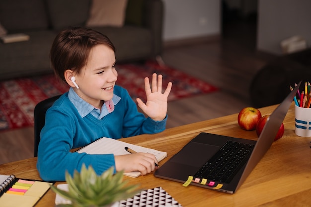 Un niño preadolescente usa una computadora portátil para hacer clases en línea, saludando a la maestra