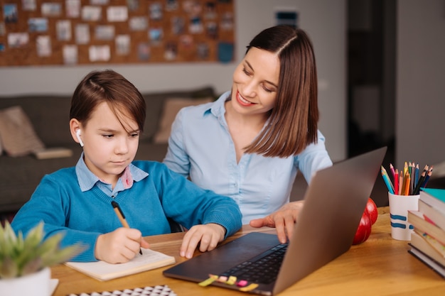 Un niño preadolescente usa una computadora portátil para hacer una videollamada con su maestra junto a su madre