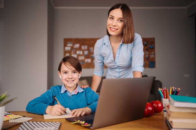 Un niño preadolescente usa una computadora portátil para hacer una videollamada con su maestra junto a su madre