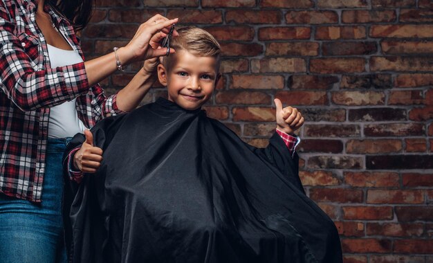 Un niño preescolar lindo y contento muestra los pulgares hacia arriba mientras se corta el pelo. El peluquero de niños con tijeras y peine está cortando a un niño pequeño en la habitación con interior de loft.