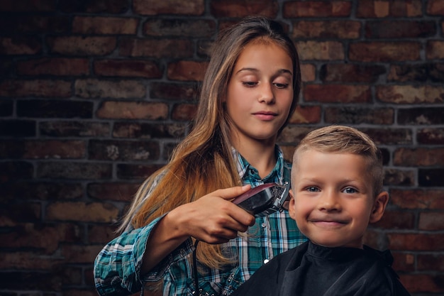 Niño preescolar sonriente cortándose el pelo. La hermana mayor corta a su hermano pequeño con una podadora contra una pared de ladrillos.