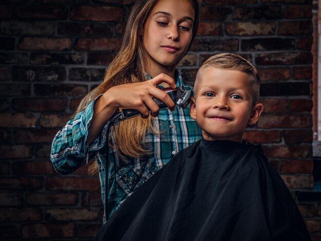 Niño preescolar sonriente cortándose el pelo. La hermana mayor corta a su hermano pequeño con una podadora contra una pared de ladrillos.