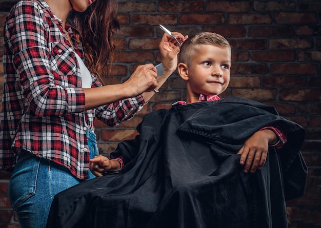 Niño preescolar sonriente cortándose el pelo. El peluquero de niños con tijeras y peine está cortando a un niño pequeño en la habitación con interior de loft.