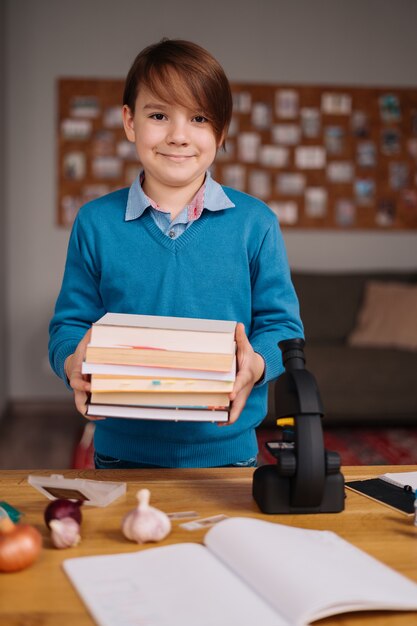 Niño de primer grado estudiando en casa, sosteniendo un montón de libros, preparándose para la lección en línea