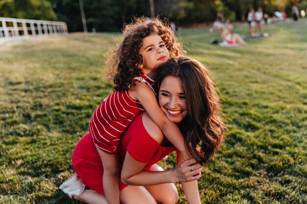 Niño relajado con pelo rizado abrazando a la madre en la naturaleza Mujer impresionante en vestido rojo jugando en el parque con su hija.