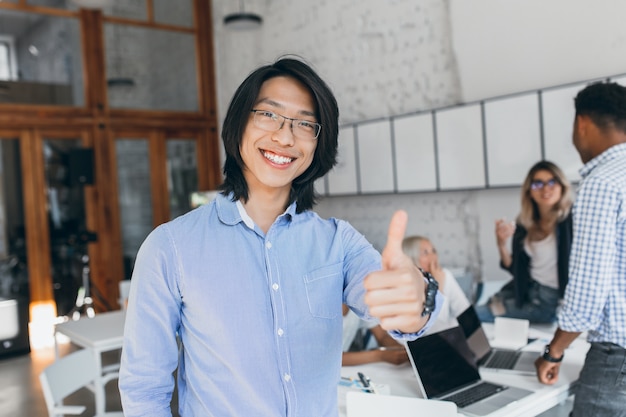 Niño riendo asiático posando con el pulgar hacia arriba al comienzo de la jornada laboral. Trabajador de oficina chino en camisa azul y gafas sonriendo con portátil.