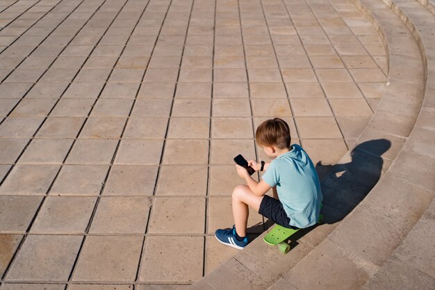 Niño sentado en las escaleras con el teléfono inteligente en la mano y penique verde viendo videos graciosos