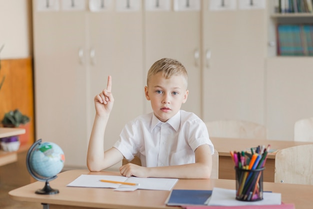 Niño sentado en el escritorio en el aula levantando la mano