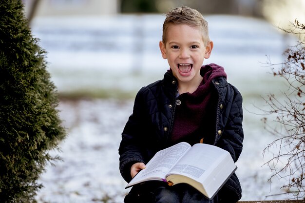 Niño sentado sobre tablas de madera y leyendo la Biblia en un jardín cubierto de nieve.