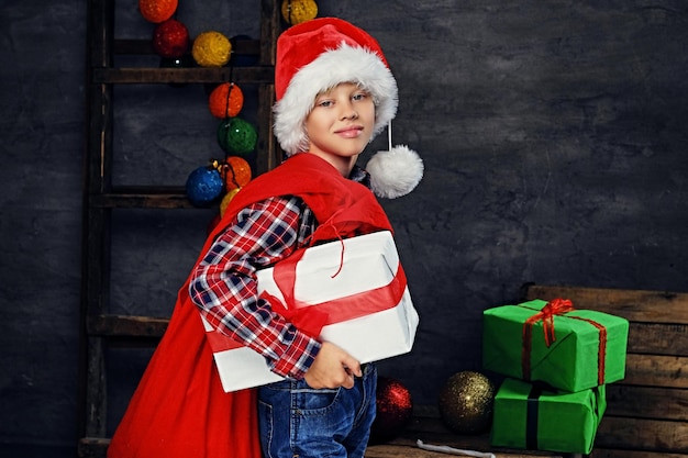 Un niño con sombrero de Papá Noel tiene una caja de regalo y una bolsa roja en el hombro.