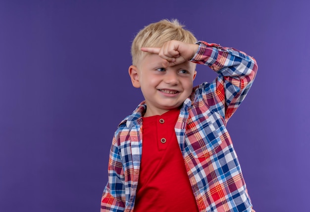 Un niño sonriente con cabello rubio vistiendo camisa a cuadros apuntando con el dedo índice a algo mientras mira de lado en una pared púrpura