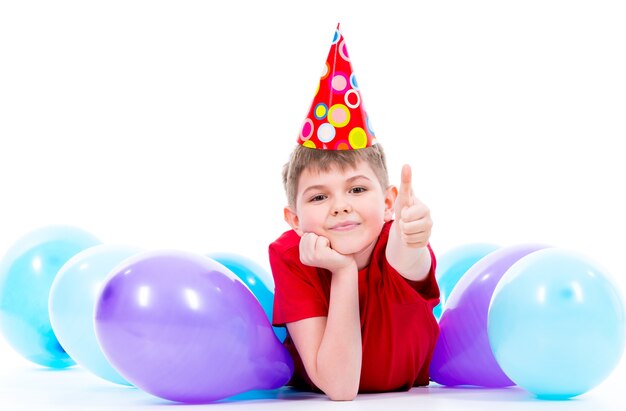 Niño sonriente feliz en camiseta roja tirado en el suelo con globos de colores y mostrando los pulgares hacia arriba - aislado en un blanco.