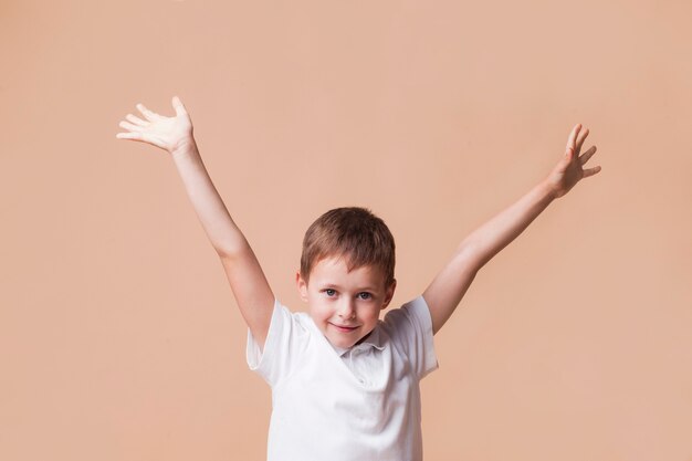 Niño sonriente inocente con la mano levantada de pie delante de fondo beige