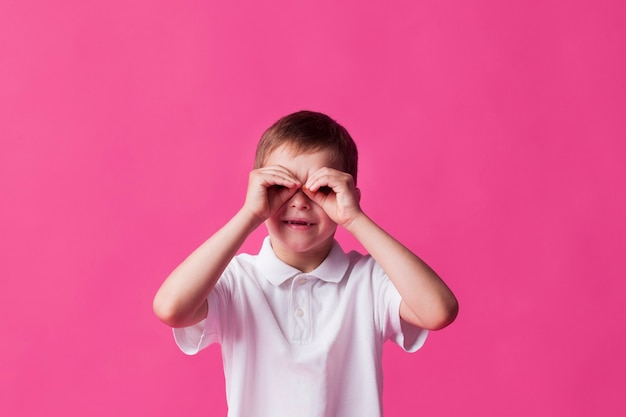 Niño sonriente mirando a través de los dedos como binoculares sobre fondo rosa