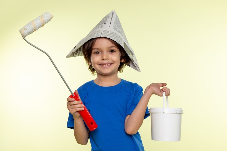 Foto gratuito niño sonriente niño en camiseta azul con pincel y pinturas en la pared blanca