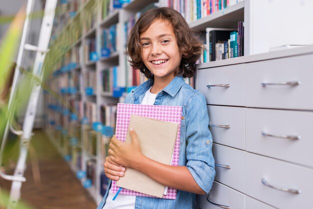 Niño sonriente sosteniendo un libro y un cuaderno en la biblioteca