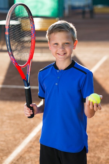 Niño sonriente sosteniendo una raqueta de tenis y una pelota