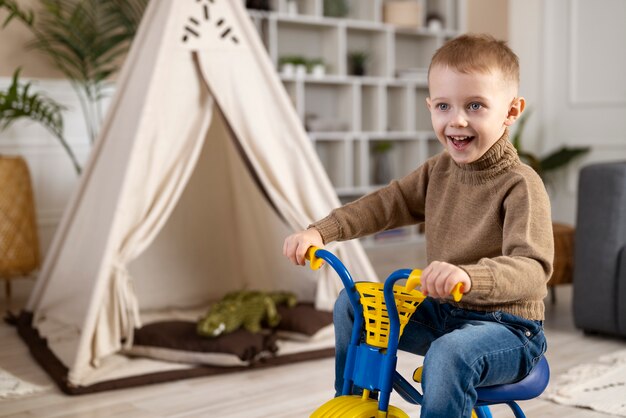 Niño sonriente de tiro completo sentado en triciclo en casa