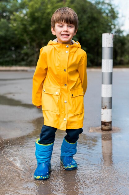 Niño con capa impermeable amarilla y botas jugando al aire libre después de  la lluvia