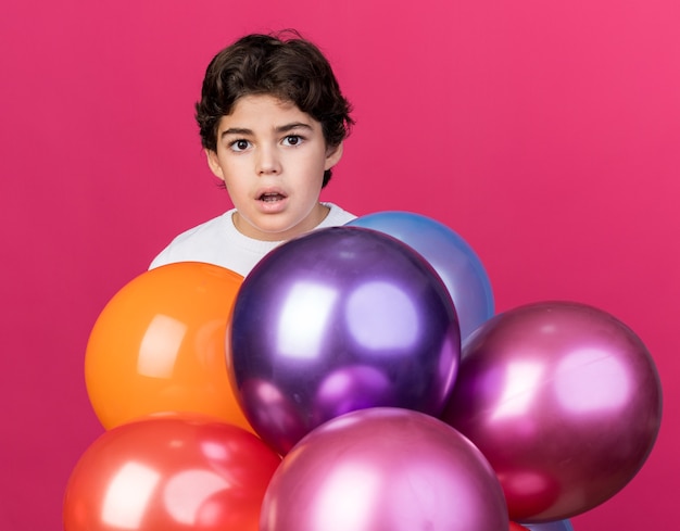 Foto gratuita niño sorprendido de pie detrás de globos aislados en la pared rosa