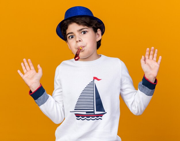 Niño sorprendido con sombrero de fiesta azul soplando silbato de fiesta extendiendo las manos aisladas en la pared naranja