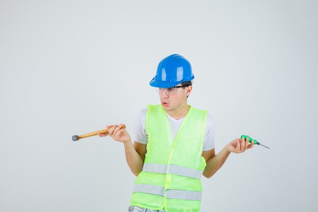 Niño sosteniendo un martillo y un destornillador en uniforme de construcción y mirando indeciso. vista frontal.