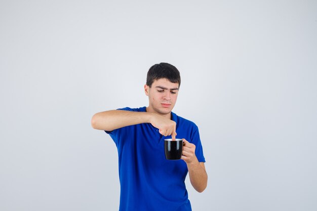 Niño sosteniendo una taza cerca de la barbilla, poniendo la mano en ella con una camiseta azul y mirando curioso, vista frontal.