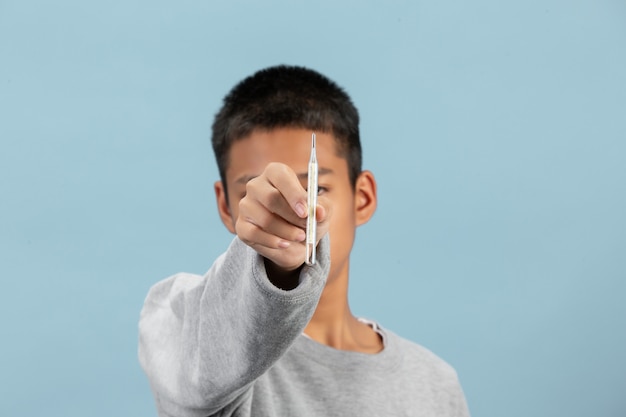 Un niño sostiene un termómetro con una camisa gris de manga larga en la pared azul.