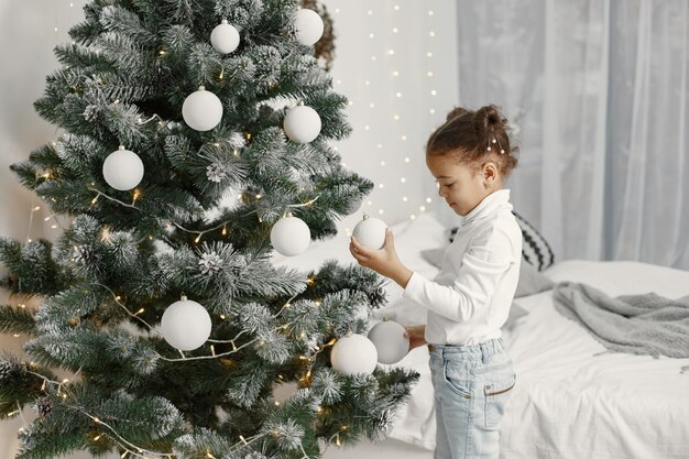 Niño con un suéter blanco. Hija de pie cerca del árbol de Navidad.