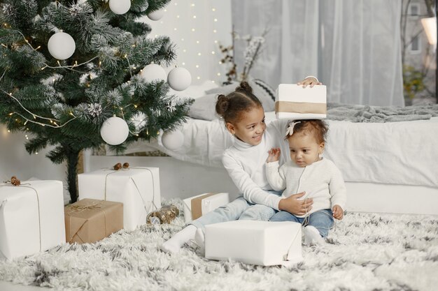 Niño con un suéter blanco. Hijas stitting cerca del árbol de Navidad. Dos hermanas en casa.