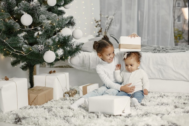 Niño con un suéter blanco. Hijas stitting cerca del árbol de Navidad. Dos hermanas en casa.