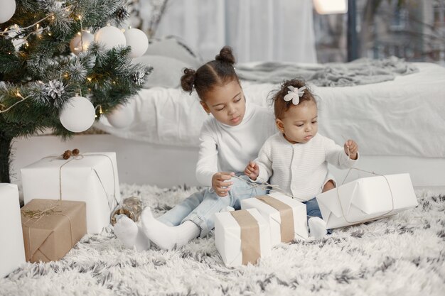 Niño con un suéter blanco. Hijas stitting cerca del árbol de Navidad. Dos hermanas en casa.