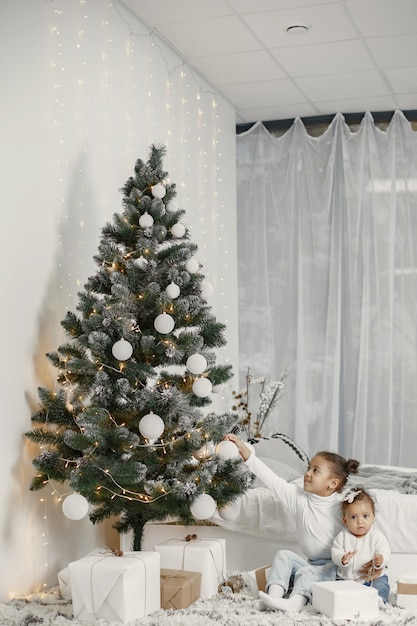 Niño con un suéter blanco. Hijas stitting cerca del árbol de Navidad. Dos hermanas en casa.