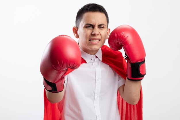 Niño superhéroe joven agresivo en capa roja con guantes de box mirando a la cámara manteniendo la mano en el aire estirando otra hacia la cámara aislada sobre fondo blanco