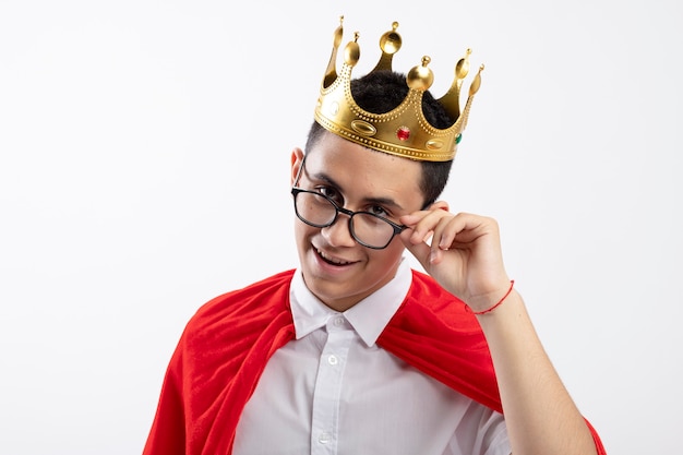 Niño superhéroe joven complacido en capa roja con gafas y corona agarrando gafas mirando a cámara aislada sobre fondo blanco