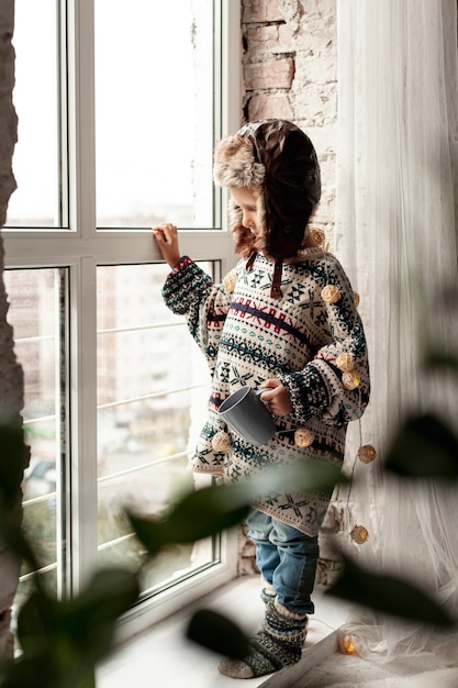 Foto gratuita niño de tiro completo con taza cerca de la ventana