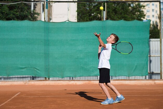 Niño tiro largo que sirve en el campo de tenis